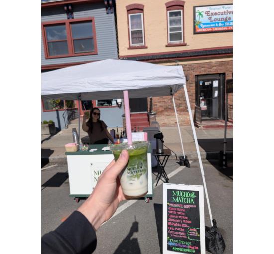 Hand holding an iced matcha latte at a Mucho Matcha outdoor street pop-up, with the chalkboard menu and cart behind