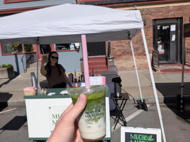 Hand holding an iced matcha latte at a Mucho Matcha outdoor street pop-up, with the chalkboard menu and cart behind